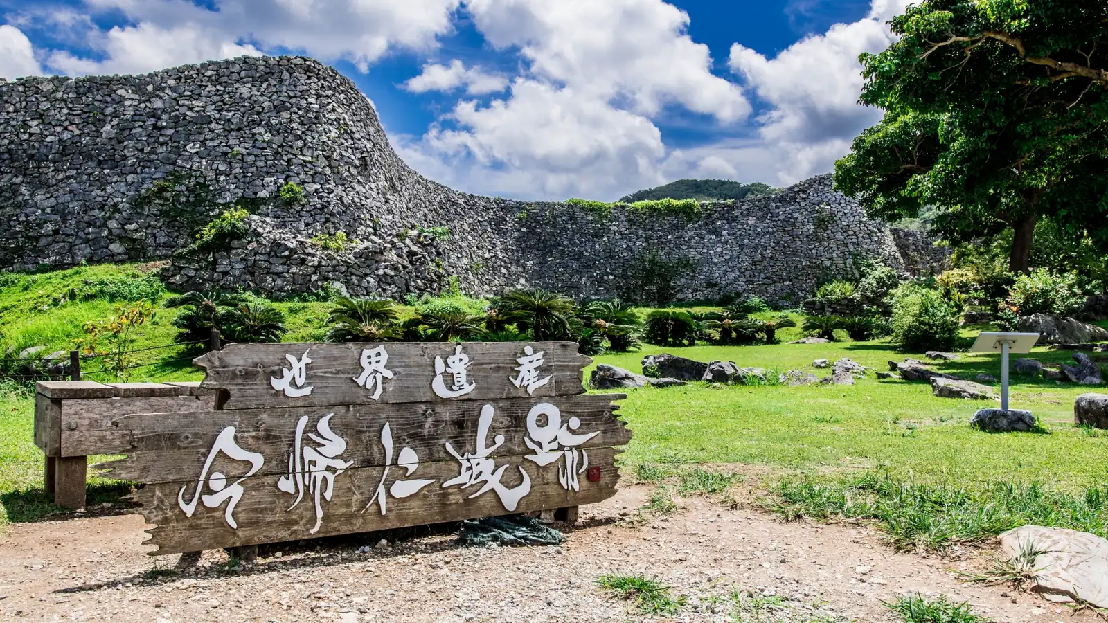 「世界遺産 今帰仁城跡」と刻まれた木の看板と、その奥にうねる石灰岩の城壁