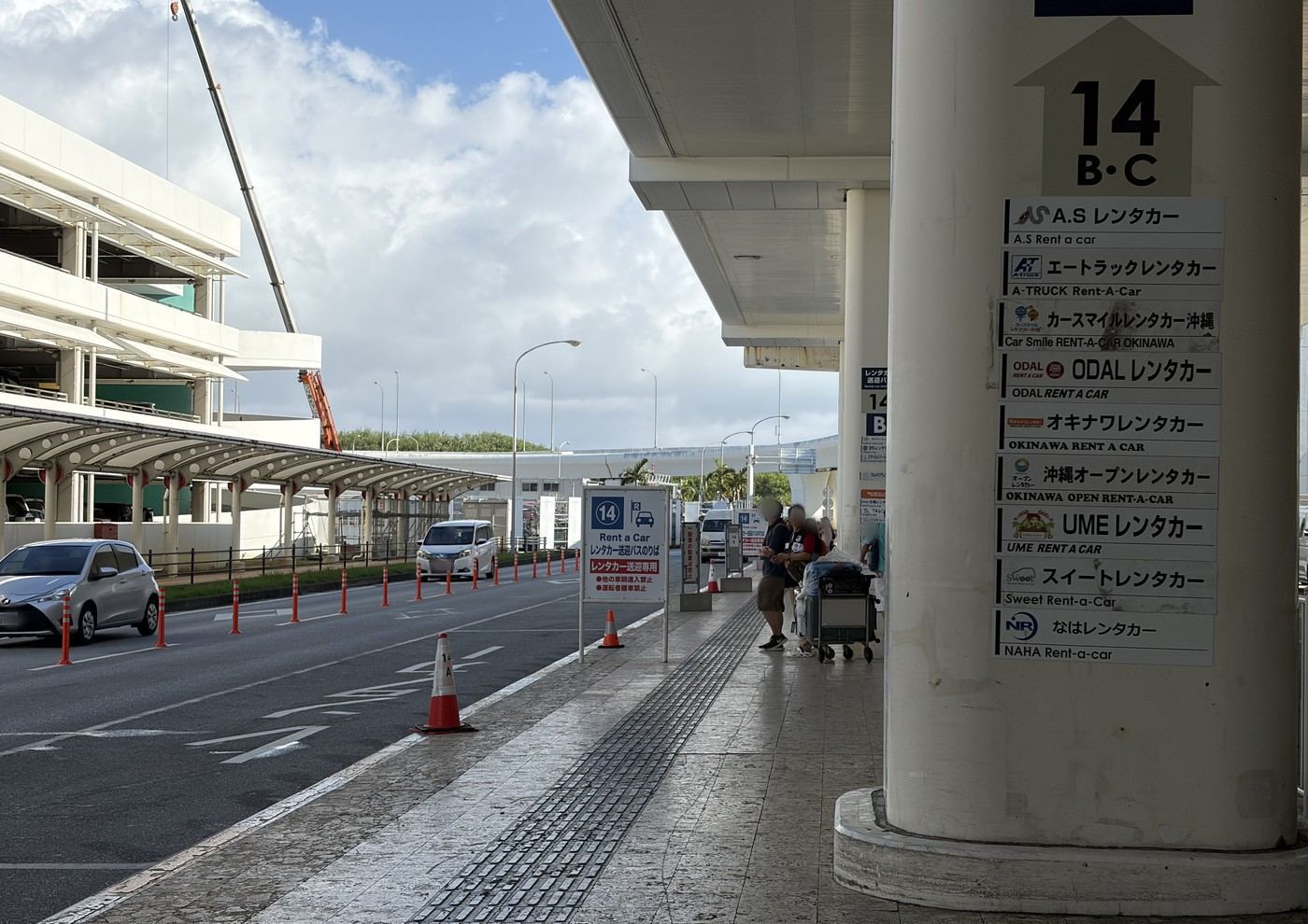 那覇空港の14番乗り場。柱に各社の看板が並び、旅行者が送迎バスを待っている